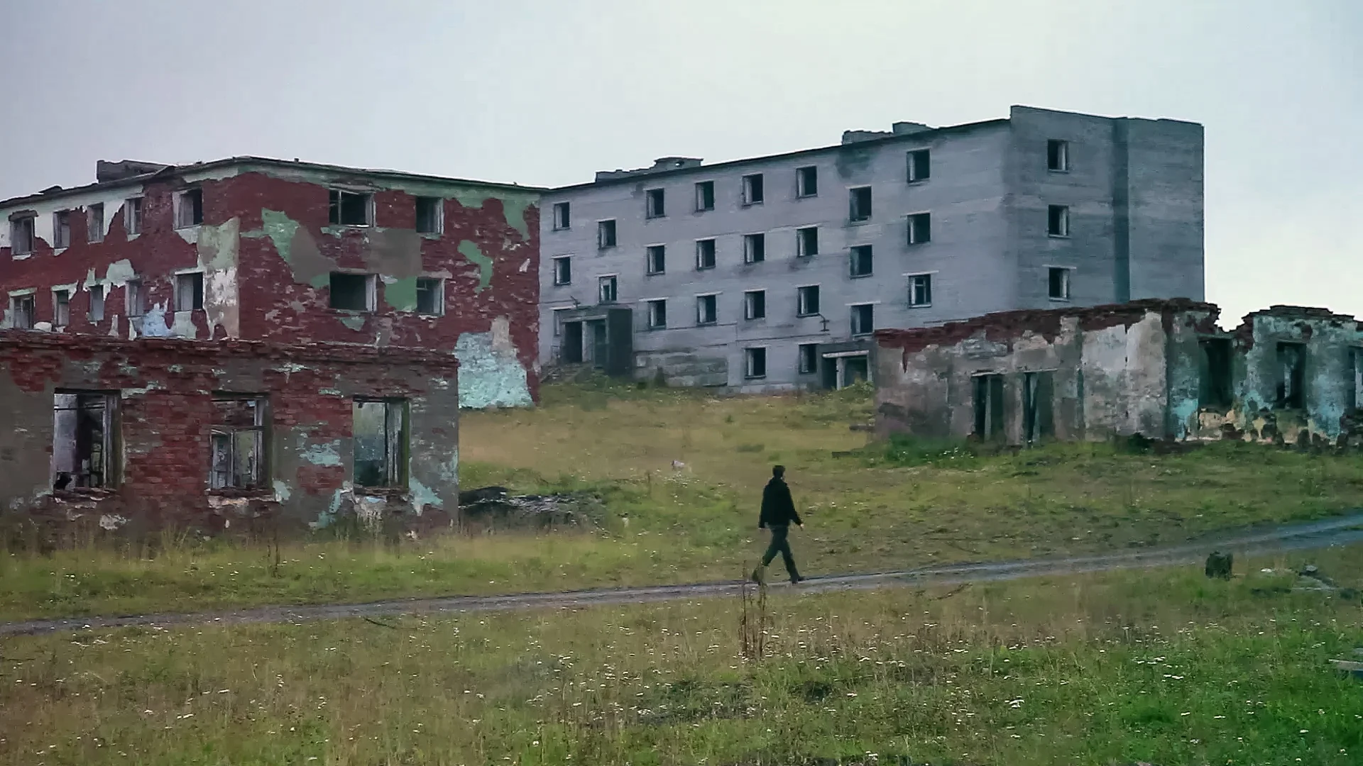A figure crossing open ground between derelict buildings
