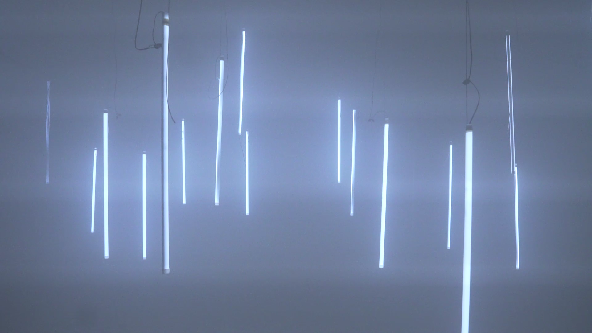 A wide installation view showing many illuminated, suspended light objects in blue-grey haze.